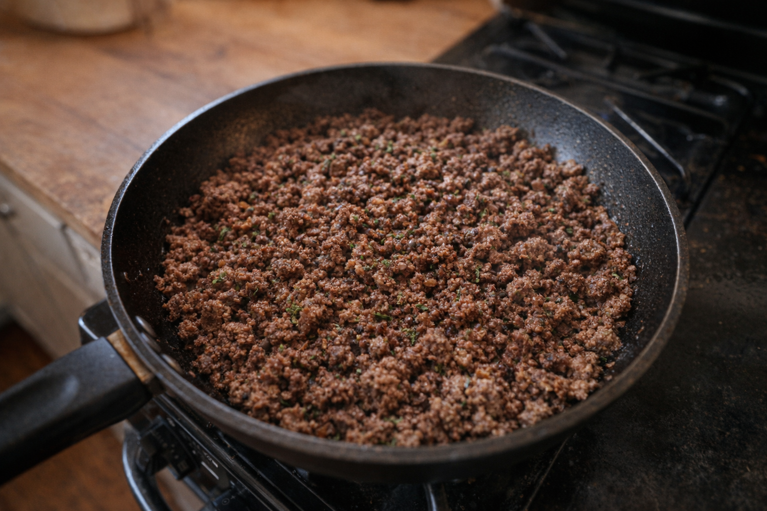 A top-down shot of browned ground beef in a skillet, evenly cooked with small bits of crispy edges, seasoned with visible herbs, warm ambient lighting